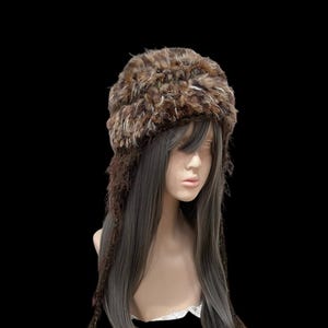 Può includere: Un cappello marrone in finta pelliccia con paraorecchie e accenti intrecciati. Il cappello è un mix di colori marrone e beige, con un aspetto strutturato. I paraorecchie si estendono verso il basso con fili intrecciati, su una testa di manichino.