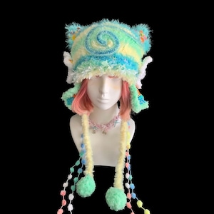 May include: A whimsical, handmade hat in pastel colors, featuring a swirl design on top and ear-like protrusions. The hat has dangling strands with pom-poms and beaded accents. The hat is displayed on a mannequin head with pink hair.
