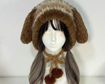 May include: A brown and beige plush hat with floppy ears, worn on a mannequin head with long gray and black hair. A matching brown knitted neckpiece with two brown pom-poms hangs below the chin. The hat is a unique, whimsical accessory.