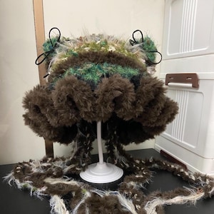 Puede incluir: Un sombrero único, hecho a mano, con un diseño caprichoso. El sombrero presenta una combinación de hilo marrón, verde y blanco, con un ala esponjosa y texturizada y detalles decorativos. El sombrero se exhibe sobre un soporte blanco, mostrando sus intrincados detalles.