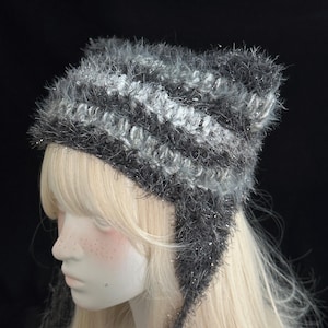 May include: A fuzzy, dark grey and silver striped beanie with earflaps. The hat is crafted from a textured, shimmery yarn that creates a soft, hairy appearance. It features alternating horizontal bands of dark charcoal and lighter silver tones.
