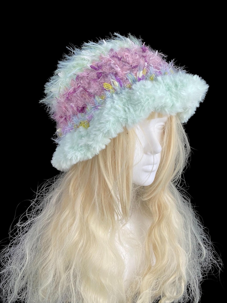 May include: A fluffy, pastel-colored hat with a wide brim. The hat features a mix of light blue, lavender, and pink yarn, creating a soft, textured appearance. The hat is displayed on a mannequin head with long, blonde hair, against a black background.