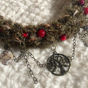 May include: A handmade necklace featuring a textured, olive-green yarn base adorned with red beads. Antique-style chains dangle from the base, with a tree-of-life pendant and circular charms.