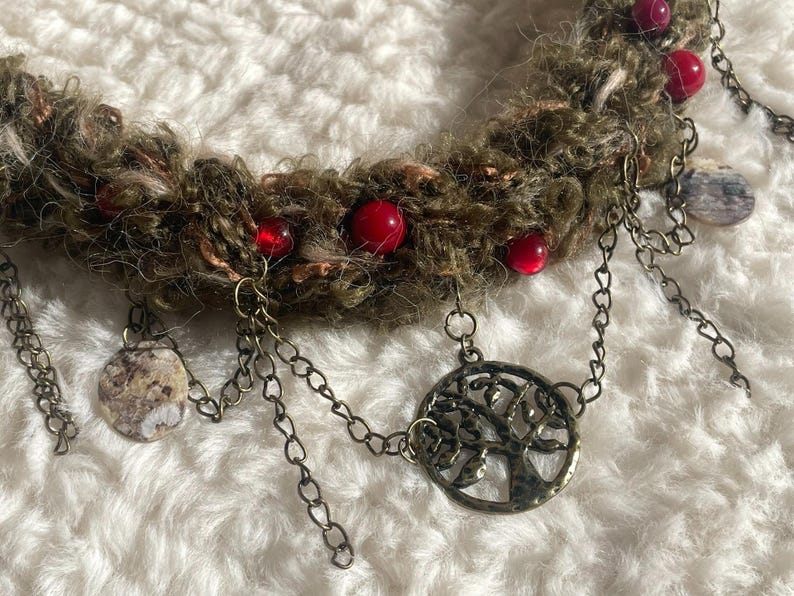 May include: A handmade necklace featuring a textured, olive-green yarn base adorned with red beads. Antique-style chains dangle from the base, with a tree-of-life pendant and circular charms.