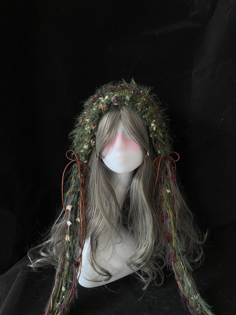 May include: A mannequin head with long, gray hair and a decorative headpiece. The headpiece is made of green and brown foliage with small flowers. Strands of the headpiece hang down, adorned with more flowers and tied with brown cords.