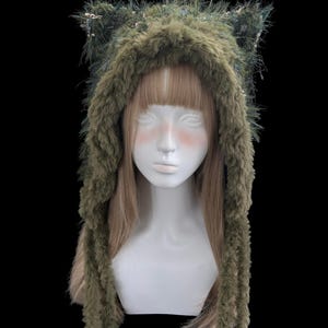 Puede incluir: Una capucha de piel sint&eacute;tica verde con orejas de gato y largas solapas para las orejas. La capucha se muestra en una cabeza de maniqu&iacute; blanca con cabello rubio. La piel es una mezcla de verde y marr&oacute;n, con peque&ntilde;os acentos rosas.