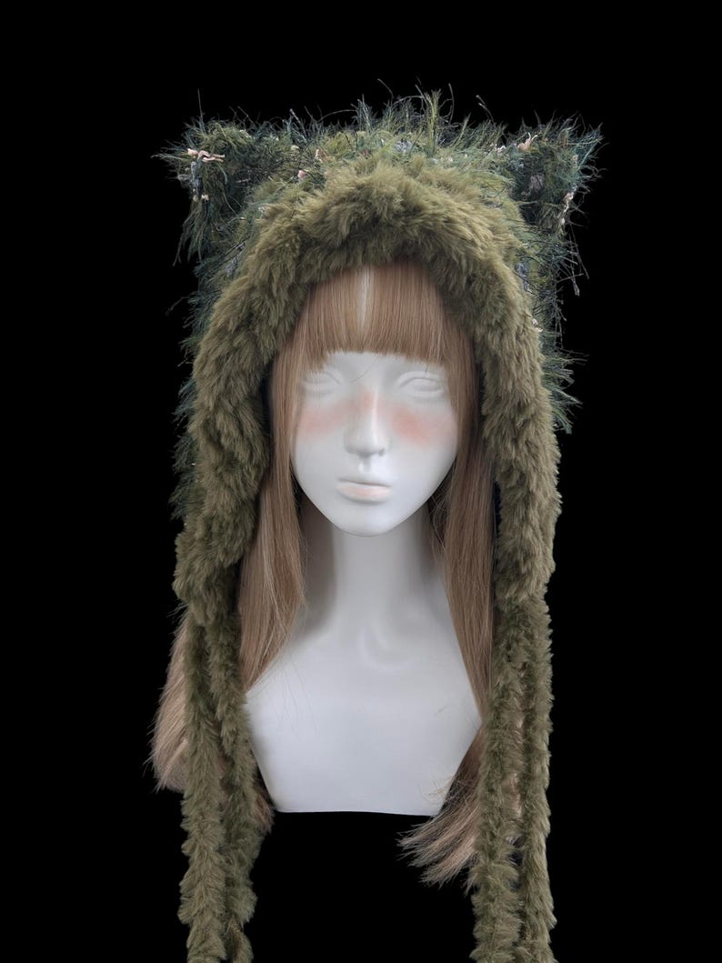 Puede incluir: Una capucha de piel sint&eacute;tica verde con orejas de gato y largas solapas para las orejas. La capucha se muestra en una cabeza de maniqu&iacute; blanca con cabello rubio. La piel es una mezcla de verde y marr&oacute;n, con peque&ntilde;os acentos rosas.