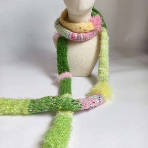 Peut inclure: Une écharpe colorée et faite à la main avec un design unique. L'écharpe présente un mélange de fils verts, jaunes, roses et bleus, avec une texture duveteuse et des éléments décoratifs. L'écharpe est présentée sur une tête de mannequin.