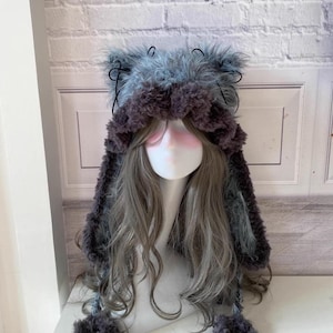 May include: A gray and brown fuzzy hat with long ear flaps and pom poms. The hat is made of soft, plush fabric and has a cute, whimsical design.