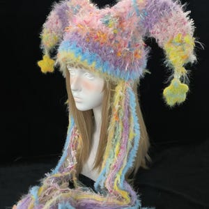 May include: A multi-coloured jester hat in pastel shades of pink, purple, yellow, and blue. The hat has long, dangling strands and star-shaped accents. The whimsical design is suitable for costume parties or festivals.