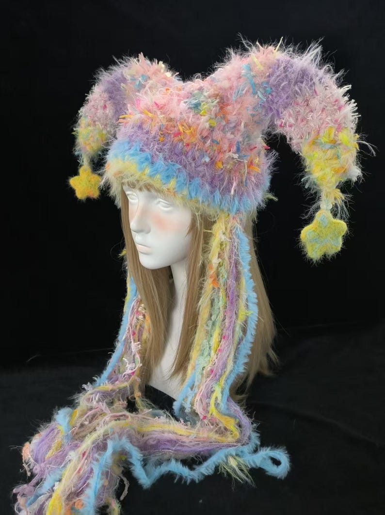 May include: A multi-coloured jester hat in pastel shades of pink, purple, yellow, and blue. The hat has long, dangling strands and star-shaped accents. The whimsical design is suitable for costume parties or festivals.