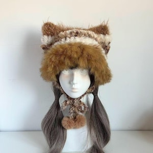 Puede incluir: Un sombrero de piel sintética marrón y blanco con orejas de gato, una bufanda a juego y dos pompones marrones. El sombrero se exhibe en una cabeza de maniquí con pelo largo gris-marrón. El sombrero tiene una textura suave y un color cálido.