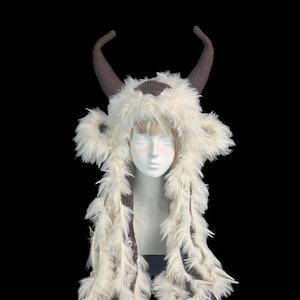 May include: A plush, cream-coloured faux fur hat featuring two large, dark brown crocheted horns on top. The hat includes long, dangling side panels of matching faux fur and a dark brown crocheted patch on the forehead, displayed on a mannequin head.