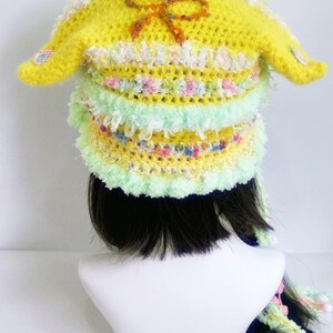 May include: A handmade yellow crochet hat with long ear flaps and a decorative bow. The hat features layers of pastel colors, including green, pink, and white, and is designed with a whimsical, playful aesthetic. The hat is a unique accessory.