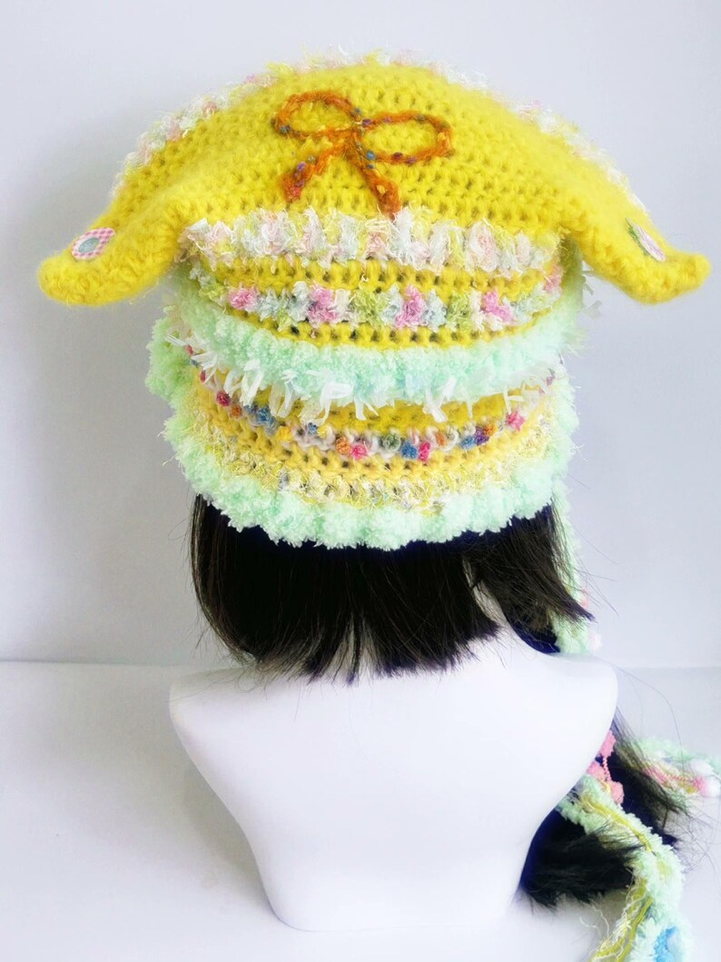 May include: A handmade yellow crochet hat with long ear flaps and a decorative bow. The hat features layers of pastel colors, including green, pink, and white, and is designed with a whimsical, playful aesthetic. The hat is a unique accessory.