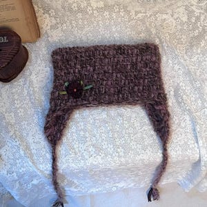 May include: A handmade, dark purple knitted hat with ear flaps. The hat features a small, decorative flower with green ribbon. The hat is made of a textured yarn and is displayed on a white lace surface.