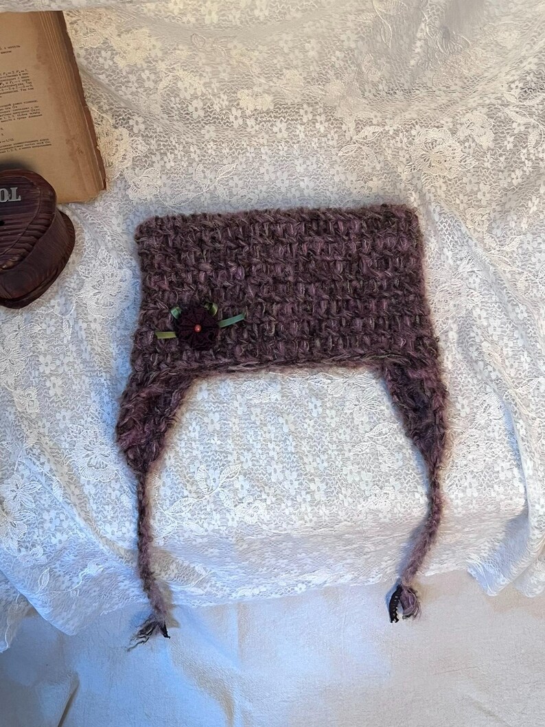 May include: A handmade, dark purple knitted hat with ear flaps. The hat features a small, decorative flower with green ribbon. The hat is made of a textured yarn and is displayed on a white lace surface.