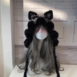 May include: Black and white furry hooded hat with long dangling ear-like strings. The hat has a fluffy texture and is designed to resemble a cat or wolf.