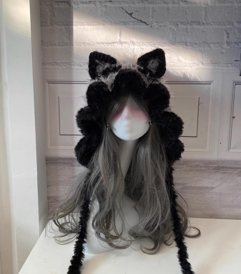 May include: Black and white furry hooded hat with long dangling ear-like strings. The hat has a fluffy texture and is designed to resemble a cat or wolf.