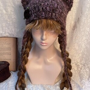 May include: A purple and pink crocheted cat ear hat with long braids. The hat features a black flower on the side and is displayed on a mannequin head. The braids are adorned with white lace and wooden beads.