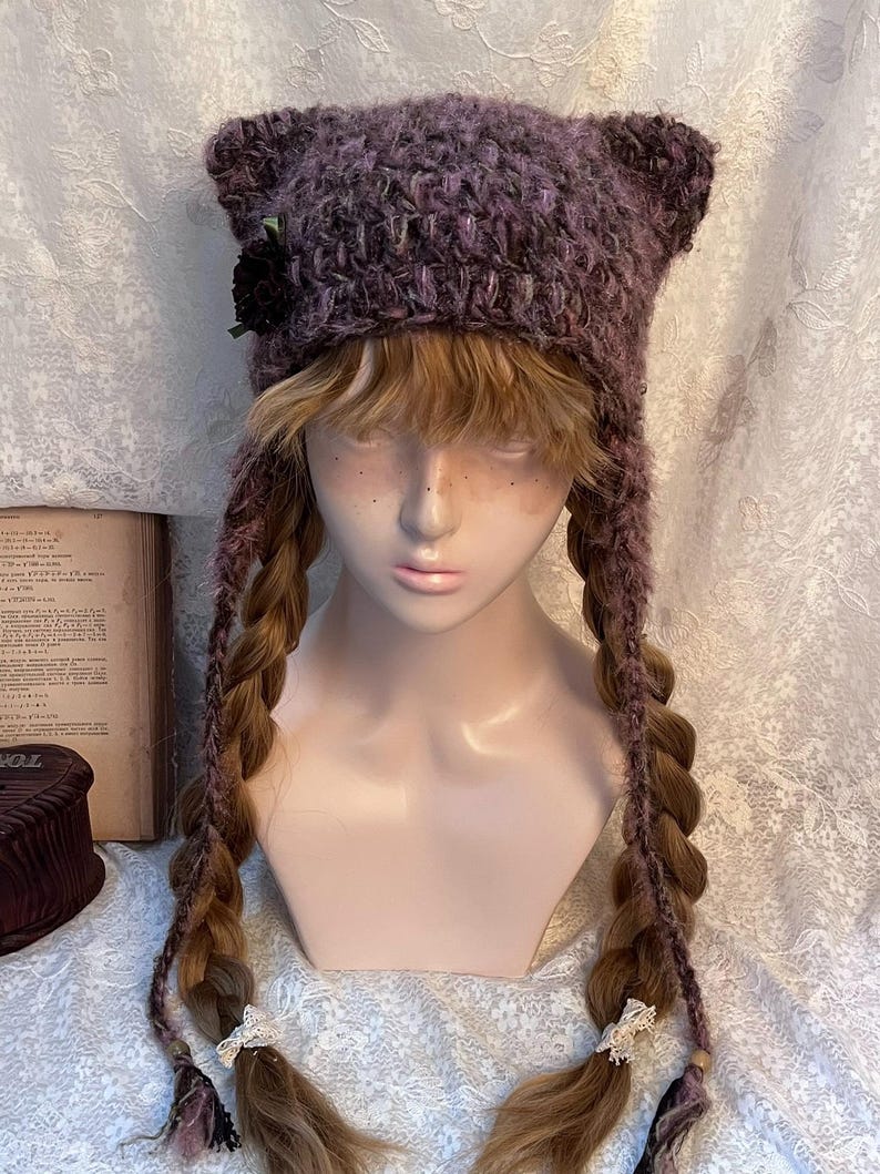 May include: A purple and pink crocheted cat ear hat with long braids. The hat features a black flower on the side and is displayed on a mannequin head. The braids are adorned with white lace and wooden beads.