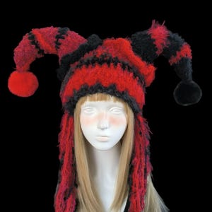 May include: A red and black jester hat with long, fringed sides and two pom-poms. The hat is made of a fuzzy material and features a zig-zag pattern. The hat is displayed on a mannequin head.