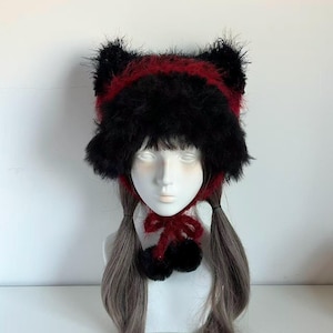 May include: A black and red cat ear hat with a fluffy texture. The hat features a red band and ear accents, with a matching bow and pom-poms. The hat is displayed on a mannequin head.