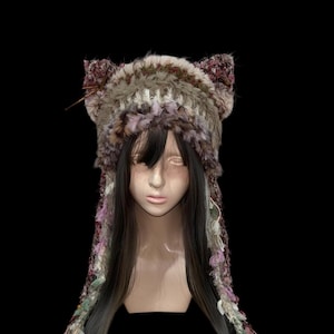 Puede incluir: Un sombrero único hecho a mano con orejas, elaborado con una mezcla de piel sintética y hilo texturizado en tonos marrón, crema y morado. El sombrero presenta largas extensiones de hilo decorativas con detalles florales, creando un diseño caprichoso.