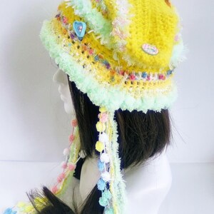 May include: A handmade, yellow crocheted hat with a mint green brim and decorative trim. The hat features colorful accents, including small pom-poms and buttons. Long, colorful strands with pom-poms hang from the hat, adding a whimsical touch.