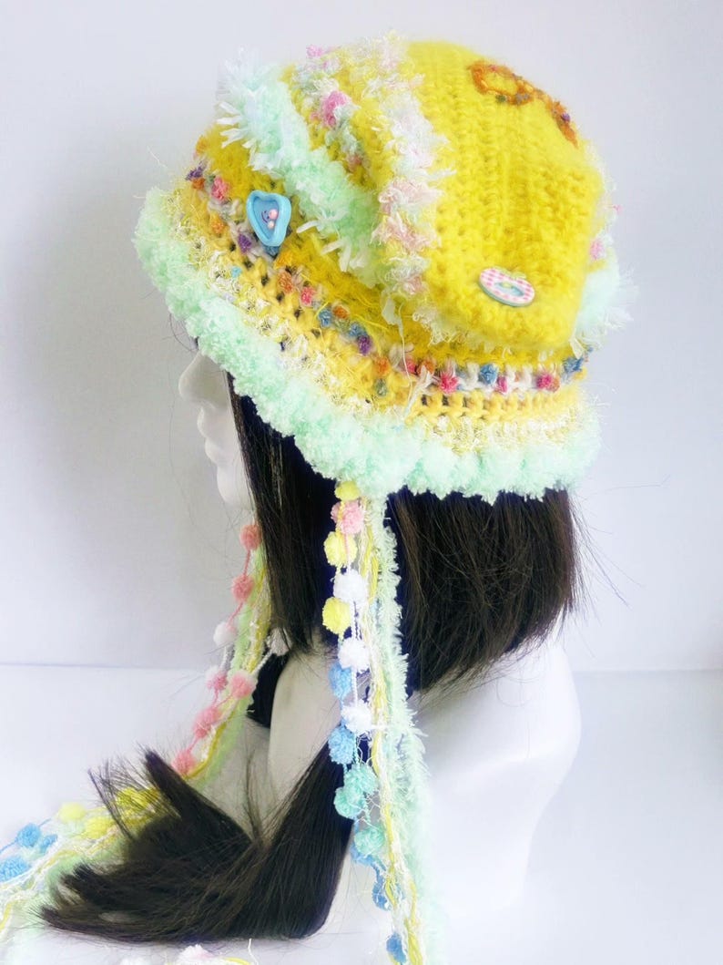 May include: A handmade, yellow crocheted hat with a mint green brim and decorative trim. The hat features colorful accents, including small pom-poms and buttons. Long, colorful strands with pom-poms hang from the hat, adding a whimsical touch.