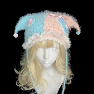 Peut inclure: Un bonnet de style bouffon en laine duveteuse, divisé en deux sections bleu clair et rose pastel. Il présente deux rabats pointus avec des pompons, une bordure blanche à volants et de longs liens pendants terminés par des formes en étoile.