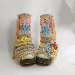 May include: A pair of platform boots with colorful, textured leg warmers. The leg warmers feature a mix of pink, yellow, and brown yarns, with blue bows and yellow fish accents. The boots have a white base and a brown sole.