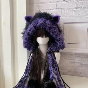 Può includere: Un cappuccio soffice in pelliccia sintetica nera e viola con orecchie da gatto integrate. Il cappuccio include lunghi pannelli laterali pendenti realizzati con lo stesso materiale testurizzato, accentuati da nastri neri e due grandi pon pon neri.