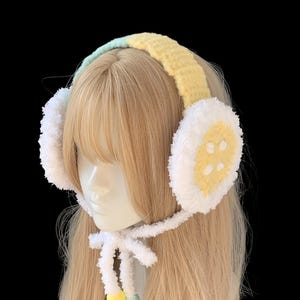 May include: Handmade earmuffs in pastel shades. The headband is yellow and light blue, with white ear covers featuring yellow button details. A white tie with yellow and light blue pom-poms completes the design. The earmuffs are designed to be worn on the head.