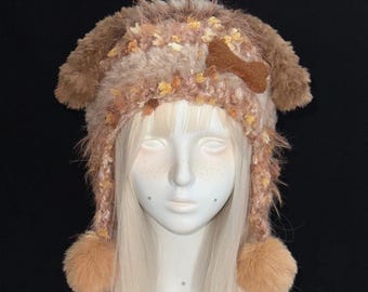 May include: A brown and tan fuzzy animal-themed hat featuring long ear flaps, two dangling pom-poms, and a brown felt bone-shaped applique on the side. The hat is crafted from textured, multi-tonal yarn with a soft, shaggy appearance throughout.