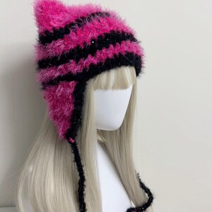K&ouml;nnte beinhalten: Eine pink-schwarz gestreifte Katzenohrm&uuml;tze mit Ohrenklappen und Bindeb&auml;ndern. Die M&uuml;tze ist aus einem flauschigen Material gefertigt und wird auf einem Schaufensterkopf mit blonden Haaren pr&auml;sentiert. Die M&uuml;tze hat ein einzigartiges, verspieltes Design.