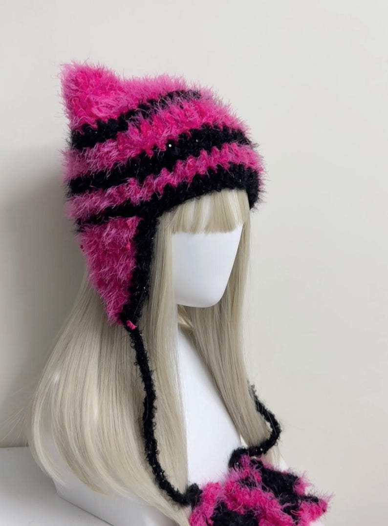 K&ouml;nnte beinhalten: Eine pink-schwarz gestreifte Katzenohrm&uuml;tze mit Ohrenklappen und Bindeb&auml;ndern. Die M&uuml;tze ist aus einem flauschigen Material gefertigt und wird auf einem Schaufensterkopf mit blonden Haaren pr&auml;sentiert. Die M&uuml;tze hat ein einzigartiges, verspieltes Design.