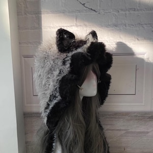 May include: A black and white furry hooded hat with cat ears. The hat is made of soft, plush fabric and has a long, flowing tail.
