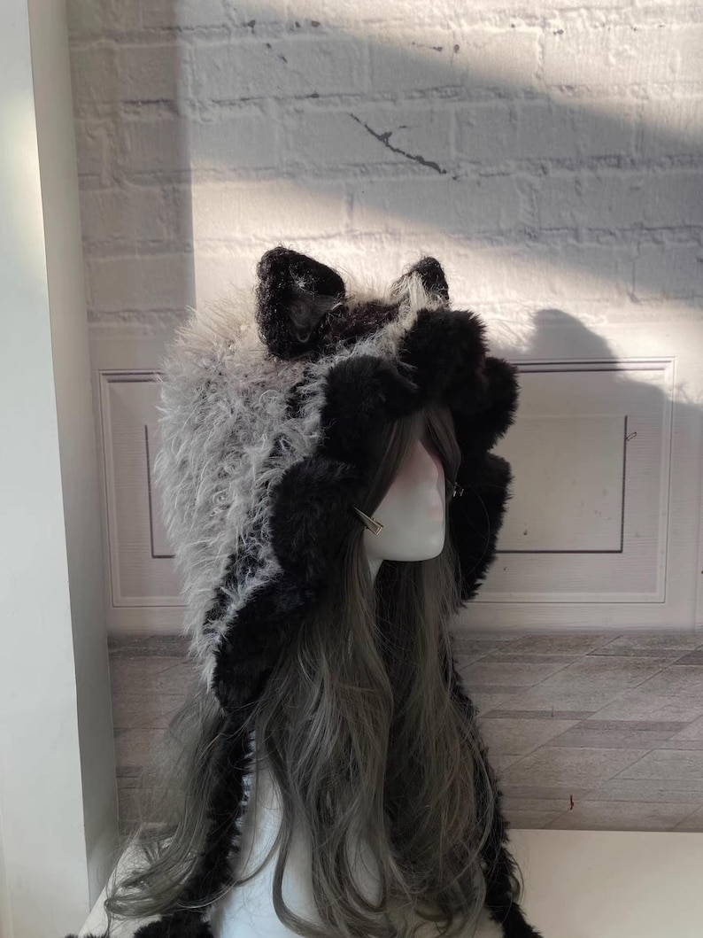 May include: A black and white furry hooded hat with cat ears. The hat is made of soft, plush fabric and has a long, flowing tail.