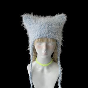 May include: A light blue fuzzy hat with pointed cat-like ears and long side tassels, displayed on a mannequin head with blonde braided hair and a bright neon yellow choker necklace against a solid black background.