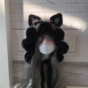 May include: A black and white furry hooded hat with cat ears. The hat has a long black fur tie.