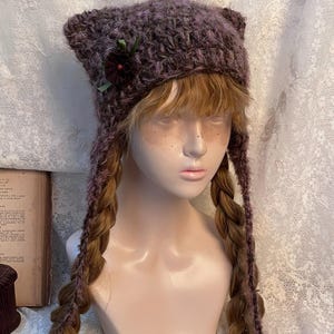 May include: A purple knit hat with ear-like points and long braided ties. The hat is decorated with a dark red flower and green ribbon. The hat is displayed on a mannequin head with long, braided hair.