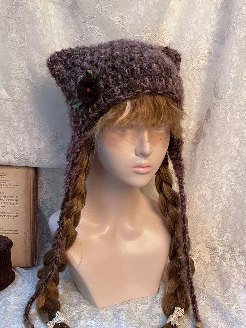 May include: A purple knit hat with ear-like points and long braided ties. The hat is decorated with a dark red flower and green ribbon. The hat is displayed on a mannequin head with long, braided hair.