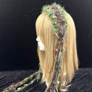 May include: A decorative headpiece with a floral design. The headpiece features a crown of green, purple, and white flowers and foliage. Long strands of the same materials hang down, tied with a brown ribbon. The headpiece is displayed on a mannequin head with blonde hair.