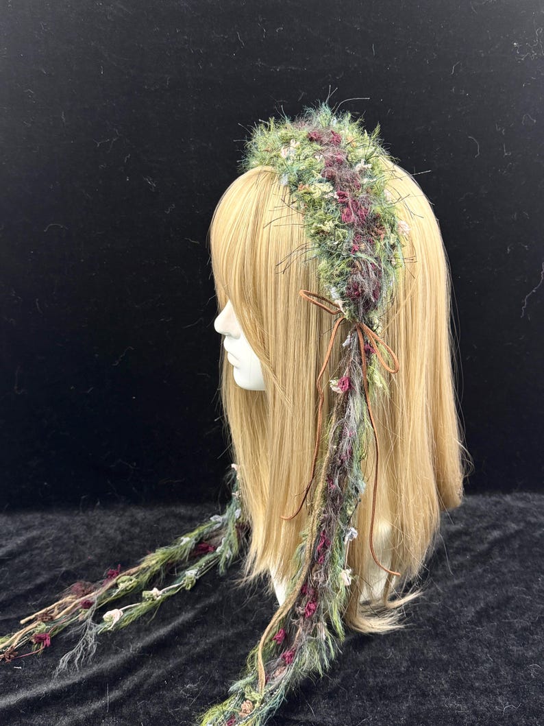 May include: A decorative headpiece with a floral design. The headpiece features a crown of green, purple, and white flowers and foliage. Long strands of the same materials hang down, tied with a brown ribbon. The headpiece is displayed on a mannequin head with blonde hair.