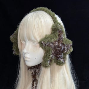 May include: A pair of fluffy green and brown star-shaped earmuffs. The ear coverings are star-shaped and have a brown and white speckled pattern. The earmuffs are connected by a matching headband made of a soft, textured material.