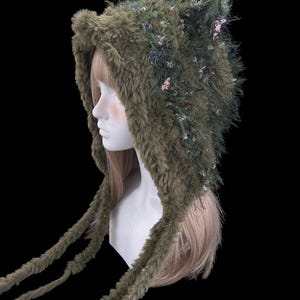 Puede incluir: Una capucha de piel sint&eacute;tica verde oliva con detalles de orejas y lazos largos. La capucha est&aacute; adornada con peque&ntilde;as flores y follaje, dando un aspecto de bosque. La capucha se muestra en una cabeza de maniqu&iacute;.