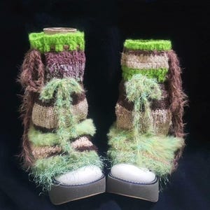 May include: A pair of platform shoes with knitted leg warmers. The leg warmers feature a color scheme of green, brown, and purple. The shoes have white soles and gray platforms. The leg warmers have a fuzzy, textured appearance.