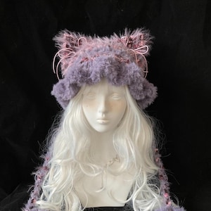 May include: A whimsical, fluffy hat in shades of lavender and pink, featuring cat ear accents. The hat is made of a soft, textured material and is designed with a unique, artistic flair. The hat is a statement piece, perfect for cosplay or costume events.