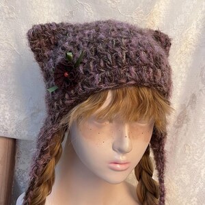 May include: A handmade, purple cat ear hat with ear flaps. The hat is adorned with a dark red flower and green ribbon. The hat is made of a textured, fuzzy yarn. The hat is designed to be worn on the head.